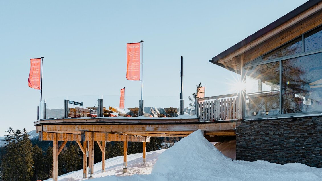 Ein modernes Restaurant auf einem Berg mit einer Holzterrasse und weißen Schneeverwehungen. Die Sonne scheint durch die Fenster, während orangefarbene Fahnen wehen. | © Knapplhütte