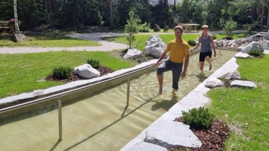 Zwei Personen gehen im Wasser eines Gehwegs durch eine grüne Parklandschaft. Umgeben von Pflanzen und Steinen strahlen die beiden Freude aus. | © Marktgemeinde Gröbming