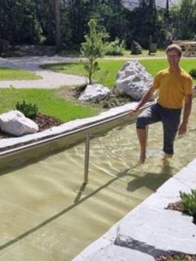 Zwei Personen gehen im Wasser eines Gehwegs durch eine grüne Parklandschaft. Umgeben von Pflanzen und Steinen strahlen die beiden Freude aus. | © Marktgemeinde Gröbming