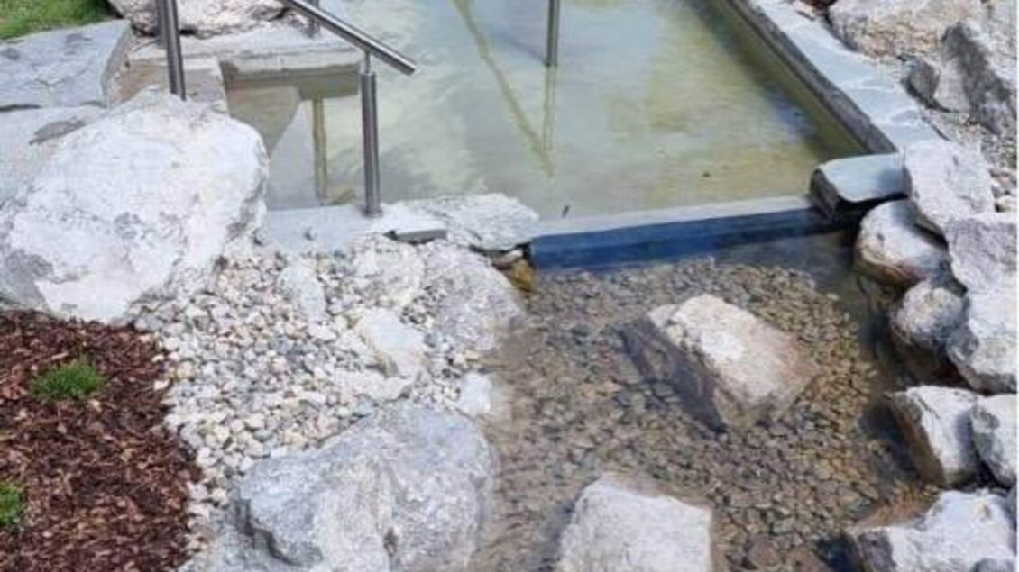 Ein natürlicher Bach mit fließendem Wasser und großen Steinen. Auf der Seite steht eine kleine Holzbank in einer gepflegten Grünanlage. | © Marktgemeinde Gröbming