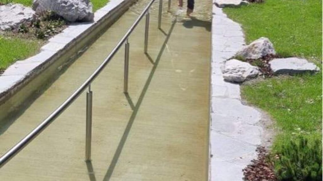 Ein ruhiger Garten mit einem Wasserlauf und gepflegtem Grün. Zwei Personen sind unterwegs, um das entspannende Ambiente zu genießen. | © Marktgemeinde Gröbming