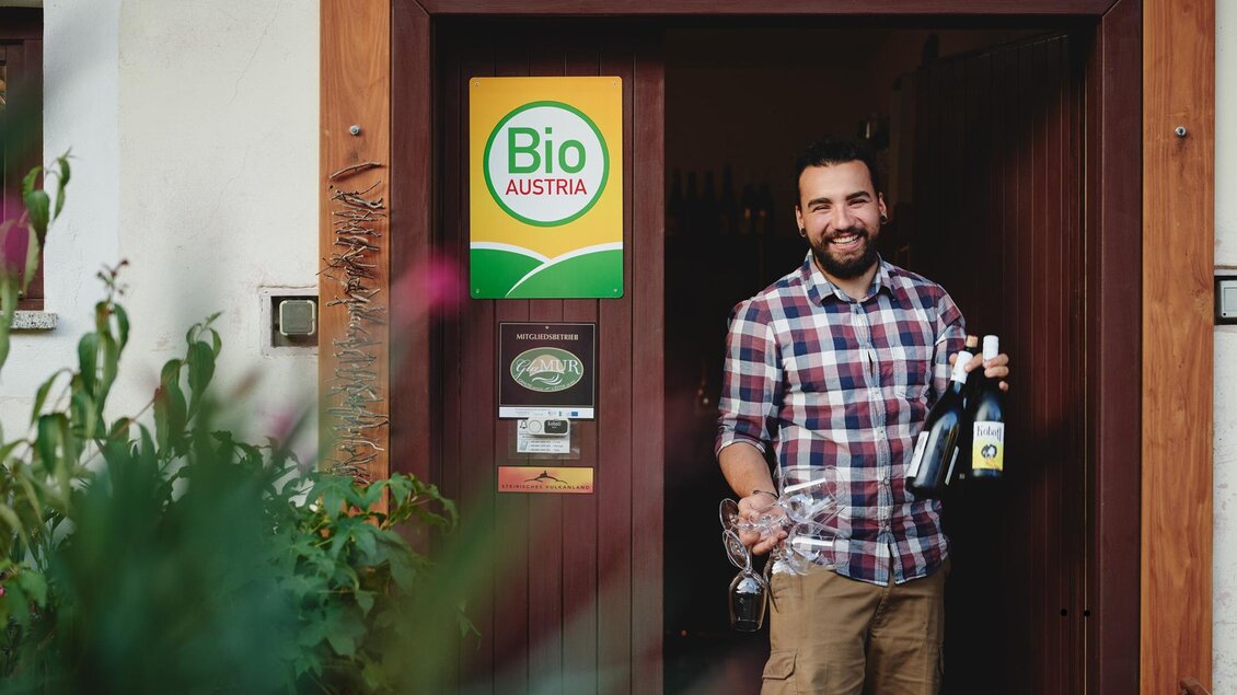 Ein Mann steht vor einem Eingang, hält zwei Flaschen Wein und ein Glas in der Hand. Im Hintergrund ist ein Schild mit der Aufschrift "Bio Austria" zu sehen. | © Kobatl