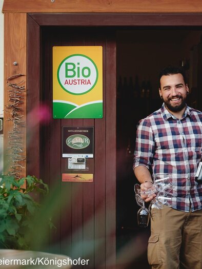 Ein Mann steht vor einem Eingang, hält zwei Flaschen Wein und ein Glas in der Hand. Im Hintergrund ist ein Schild mit der Aufschrift "Bio Austria" zu sehen. | © Kobatl