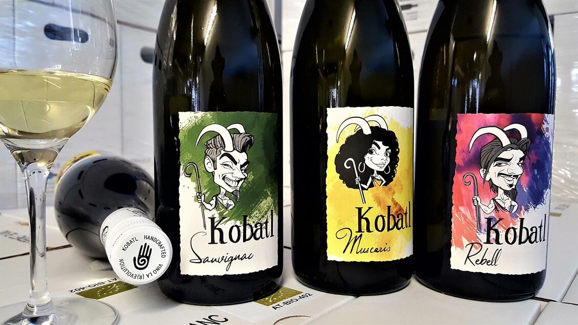 Drei Weinflaschen mit farbenfrohen Etiketten stehen auf einem Tisch. Ein Glas Weißwein sorgt für eine einladende Atmosphäre. | © Kobatl