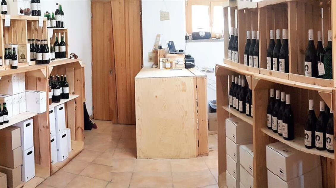 Ein Innenraum mit Holzregalen, die mit Flaschen gefüllt sind. Es gibt einen mittigen Serviertisch und eine gemütliche Atmosphäre. | © Kobatl