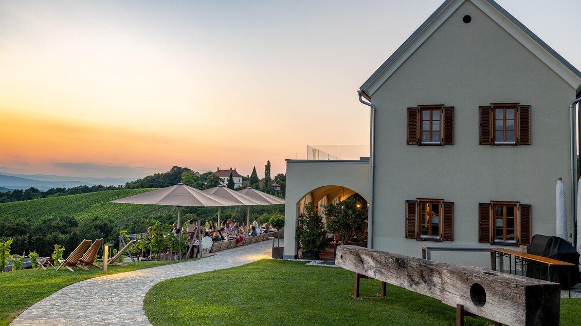 Ein schönes Haus mit Holzfenstern in einer ländlichen Umgebung. Im Hintergrund sind Weinberge und eine Terrasse mit Gästen zu sehen, während die Sonne untergeht. | © Kogel 3
