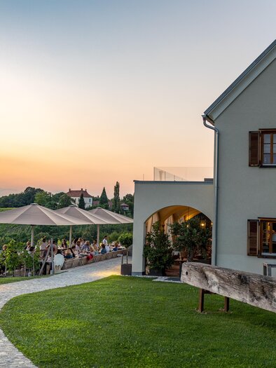 A beautiful house with wooden windows in a rural setting. In the background, vineyards and a terrace with guests can be seen, as the sun sets. | © Kogel 3
