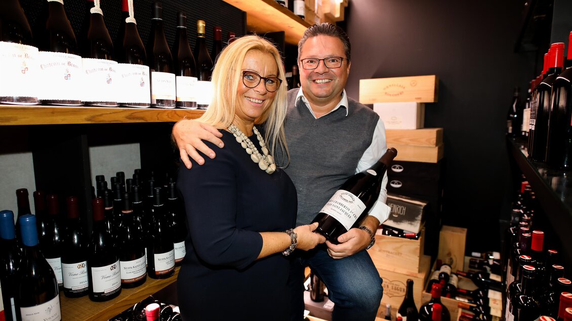 Ein Paar steht in einem Weinkeller umgeben von Weinflaschen. Sie halten eine große Flasche Wein und lächeln in die Kamera. | © Kogel 3