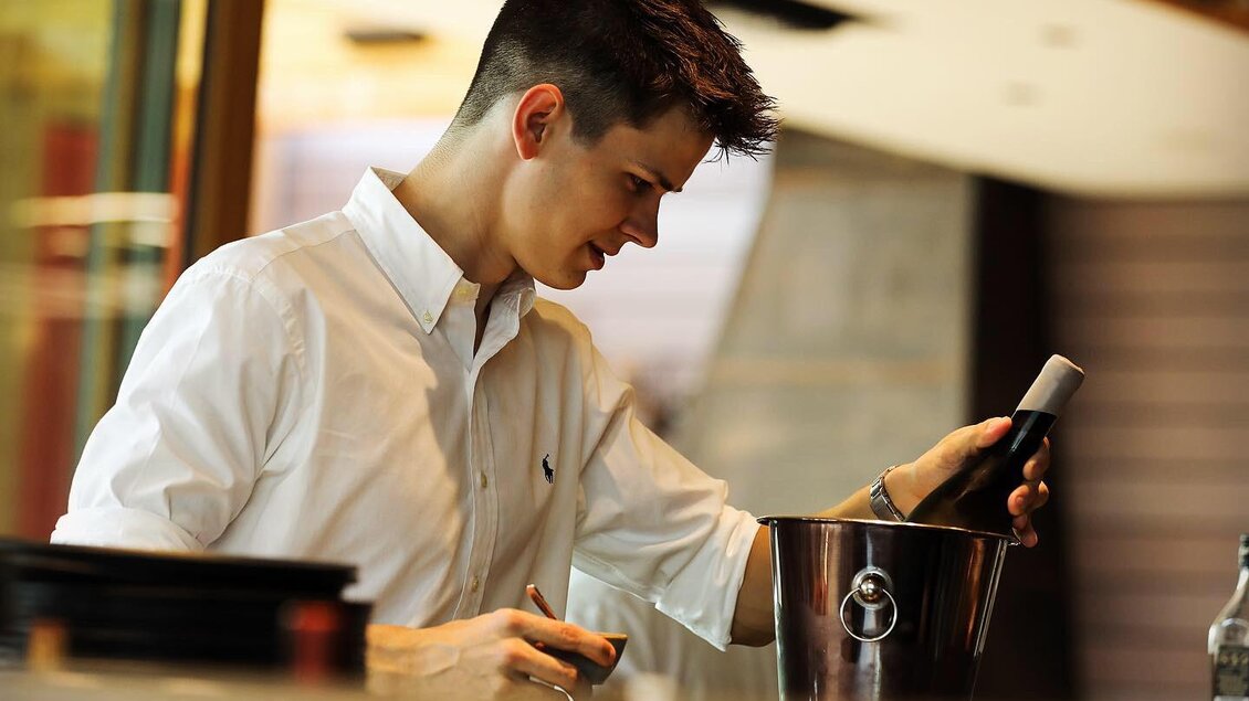 Ein junger Mann in einem weißen Hemd öffnet eine Flasche Sekt in einem Eiskübel. Die Umgebung ist modern und elegant gestaltet. | © Kogel 3