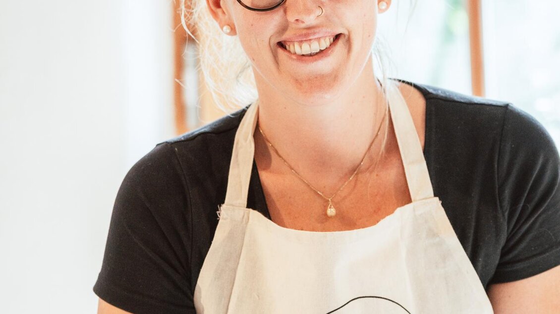 Eine lächelnde Frau trägt eine Schürze mit dem Aufdruck "Pottery Treat". Sie steht in einem hellen Raum mit großen Fenstern.