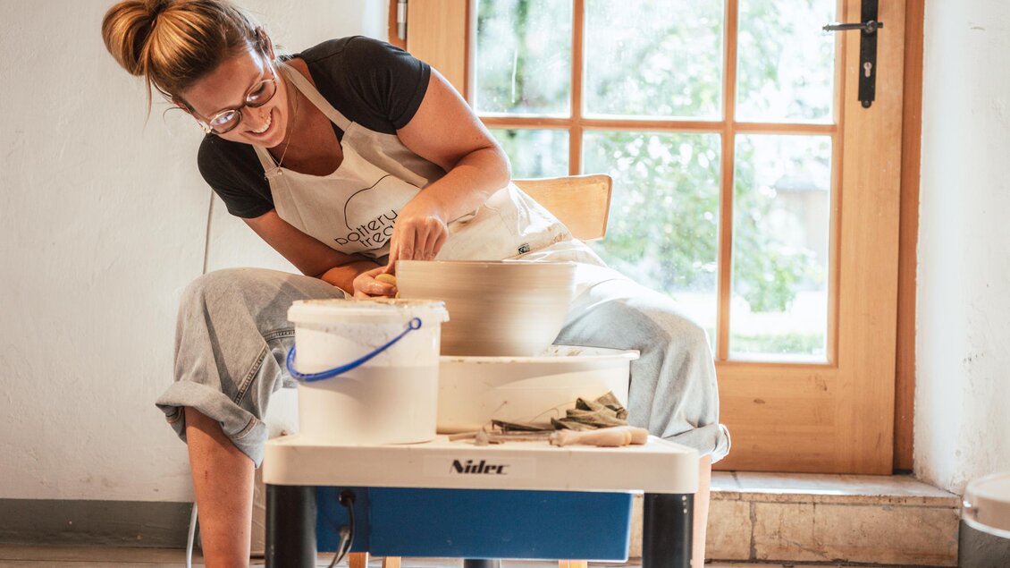 Eine Töpferin arbeitet an einer Töpferscheibe in einem hellen Raum. Sie trägt eine Schürze und hat Spaß am kreativen Prozess.