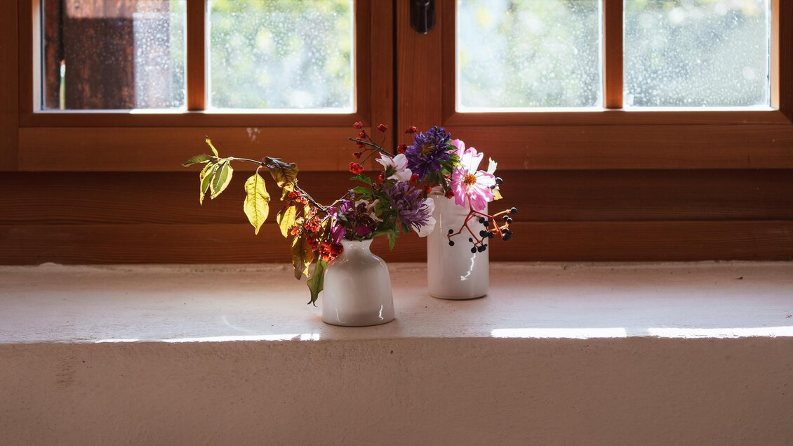 Ein heller Fensterplatz mit zwei Vasen, die farbenfrohe Blumen enthalten. Die Sonne scheint durch das Fenster und schafft eine warme Atmosphäre.