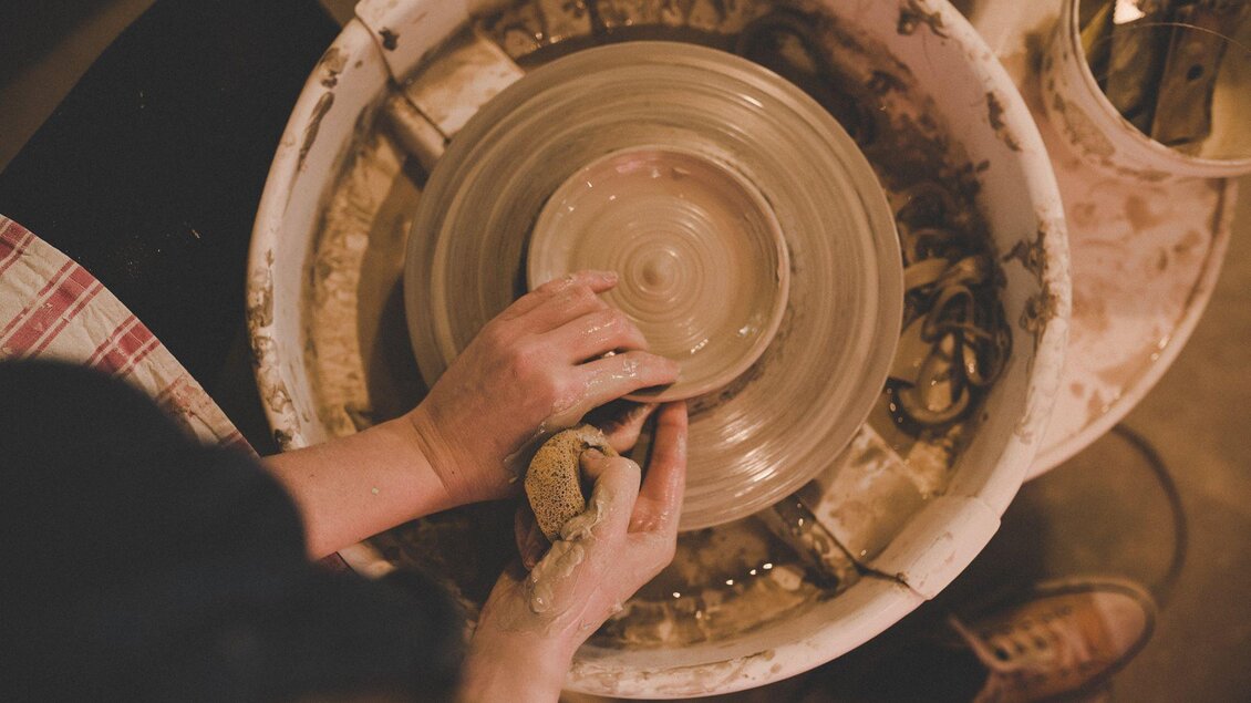 Eine Person formt Ton auf einer Töpferscheibe. Der Arbeitsplatz ist mit Lehm und Töpferwerkzeugen ausgestattet.
