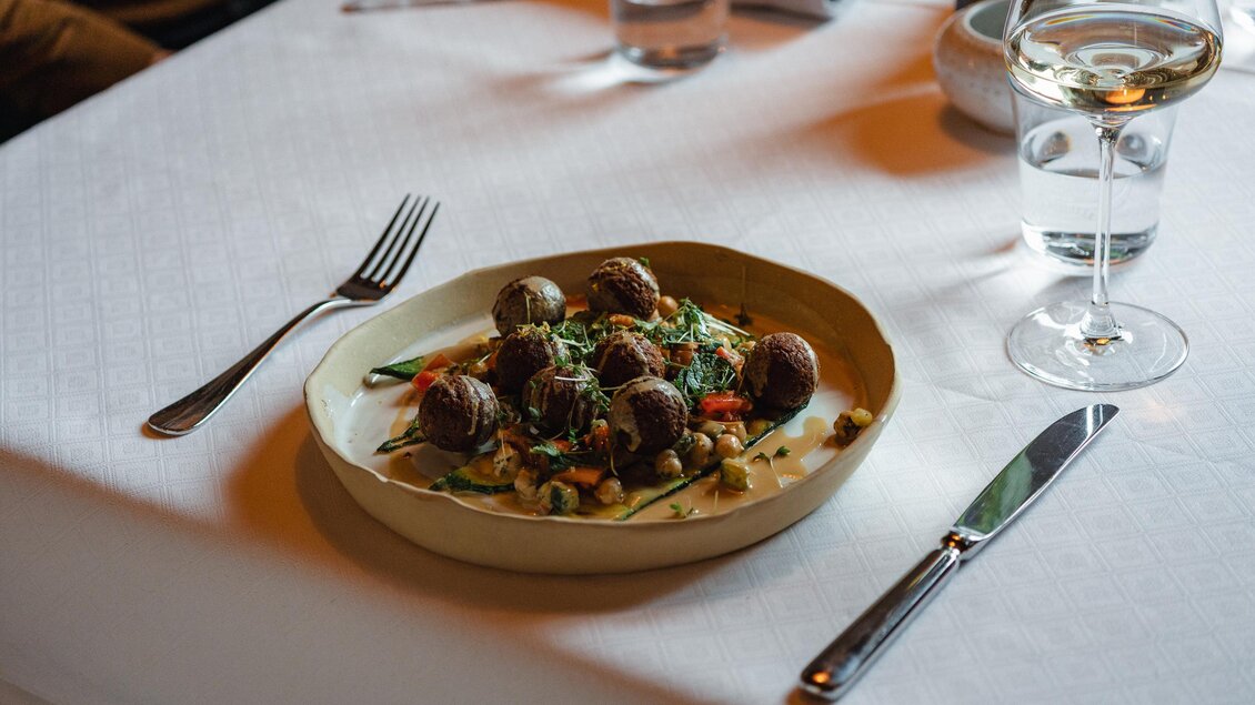 Ein Teller mit Fleischbällchen auf einem Bett aus Gemüse und Kräutern. Dazu ein Glas Wasser und Besteck auf einem weißen Tisch.