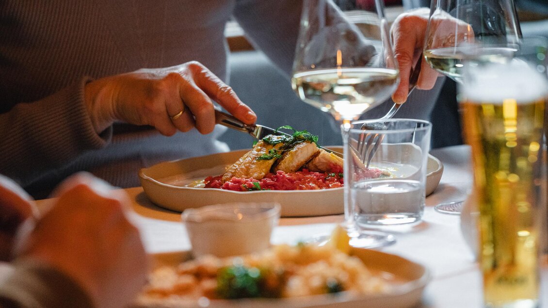Ein gedeckter Tisch mit einem köstlichen Essen und Getränken. Eine Person schneidet das Essen, während eine andere ein Glas Wein hält.