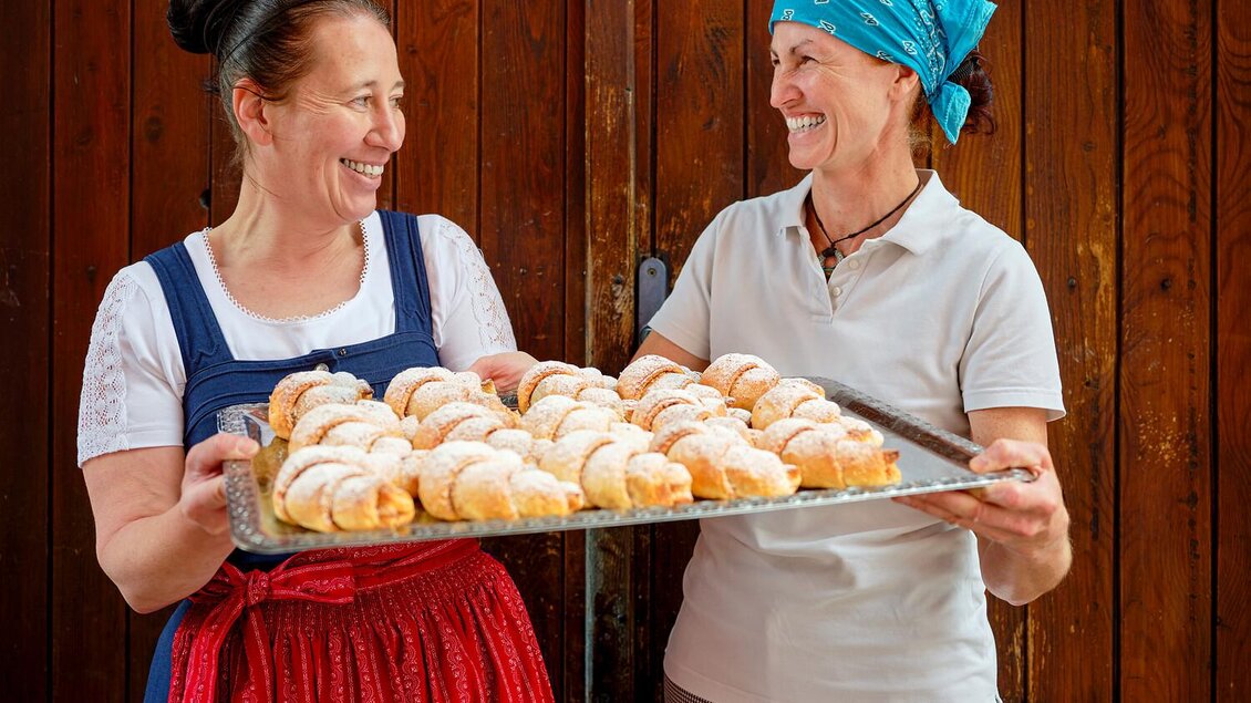 Zwei Frauen lächeln sich an und halten ein Tablett mit frischen Croissants. Der Hintergrund ist eine rustikale Holzwand. | © Susanne Enzenberger Red Bull Mediahouse