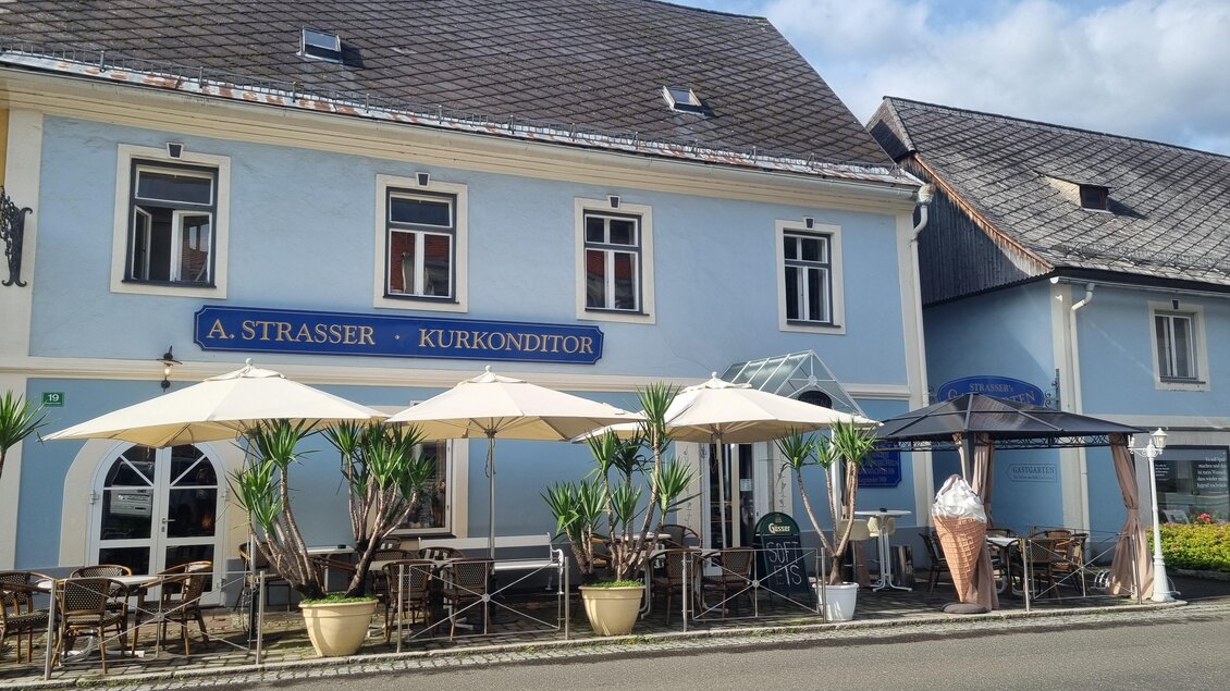 Ein Café mit einem blauen Gebäude und Tischen im Freien. Sonnenschirme und Pflanzen schaffen eine einladende Atmosphäre. | © Erlebnisregion Murtal