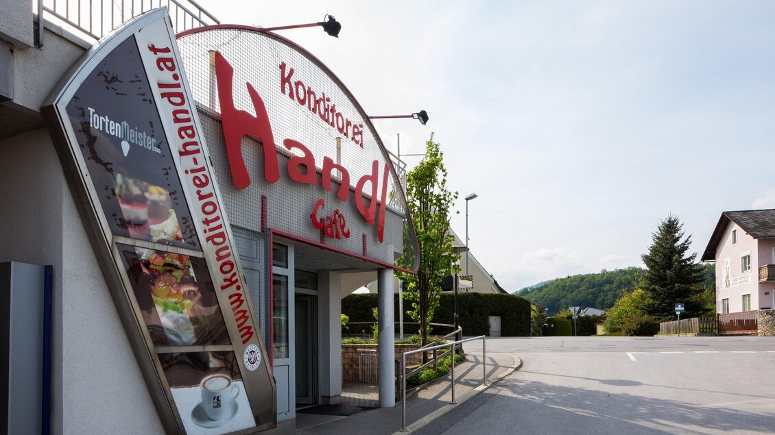 Ein einladender Eingang zu einer Konditorei mit dem Namen "Handl". Die Umgebung ist ruhig mit Bäumen und einer Straße. | © TV Region Graz - Lunghammer