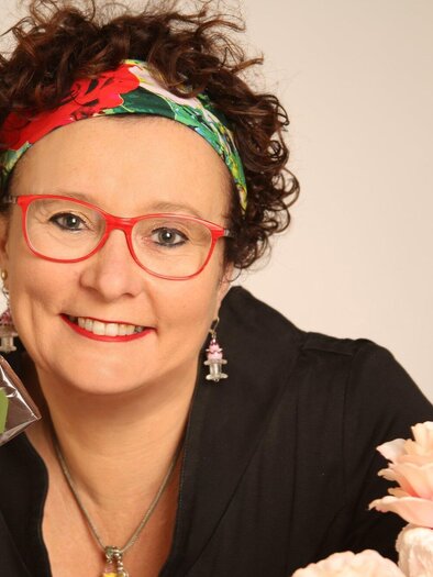 A smiling woman with curly hair and a colorful headband holds two pieces of chocolate in her hand. Next to her is a pretty cake with a flower. | © Konditorei Philipp