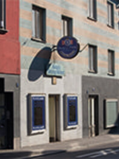 A brightly painted building on a street. There are several windows and a sign above the entrance. | © C.Stein