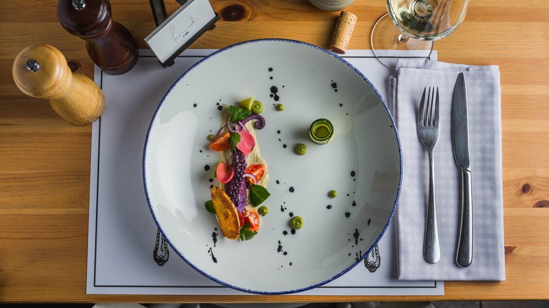 Ein kunstvoll angerichteter Teller mit buntem Gemüse und einer Hauptzutat in der Mitte. Neben dem Teller sind Besteck und ein Glas Wein auf einem Tisch platziert. | © Kornati