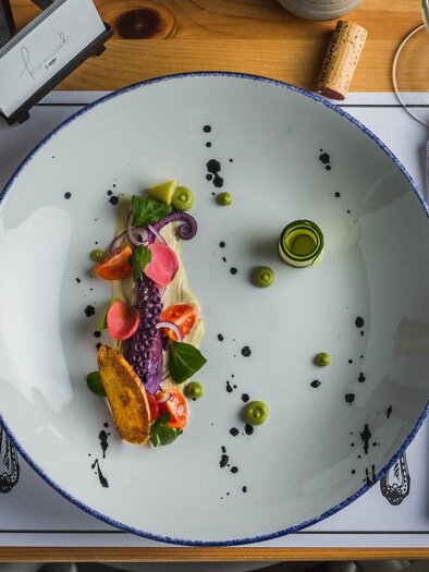 Ein kunstvoll angerichteter Teller mit buntem Gemüse und einer Hauptzutat in der Mitte. Neben dem Teller sind Besteck und ein Glas Wein auf einem Tisch platziert. | © Kornati