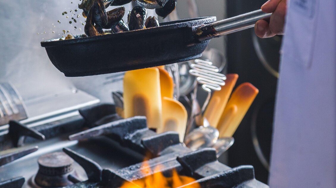 Ein Koch hebt eine Pfanne mit Muscheln über einem lodernden Herd. Küchenutensilien sind im Hintergrund sichtbar. | © Kornati