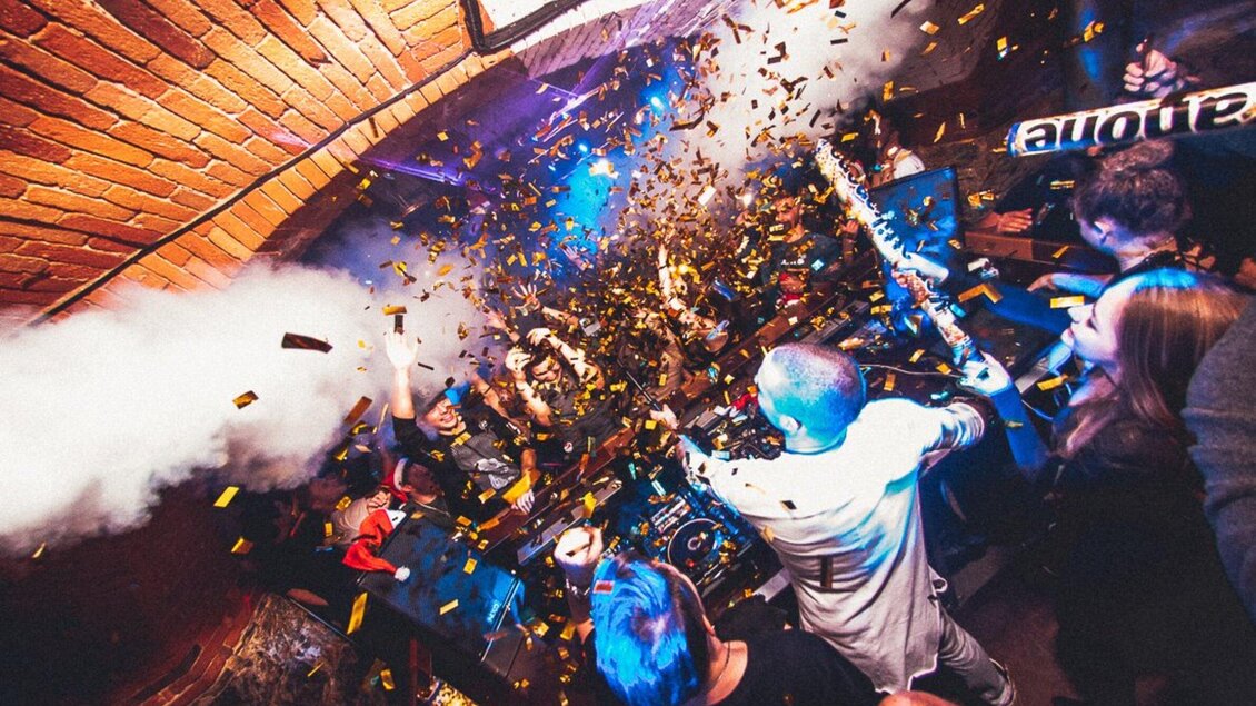 Eine ausgelassene Feier in einem Club mit tanzenden Menschen. Konfetti und Lichtstrahlen schaffen eine festliche Atmosphäre. | © Kottulinsky
