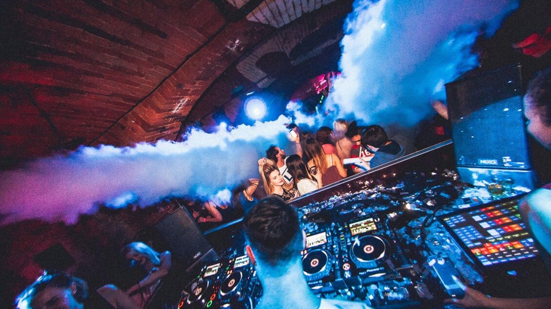 Eine lebhafte Party mit einem DJ am Mischpult und einer großen Menge von tanzenden Menschen. Bunte Lichter und Nebel verleihen der Atmosphäre eine aufregende Stimmung. | © Kottulinsky
