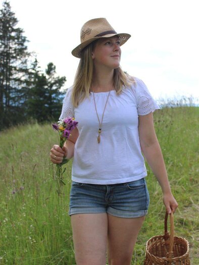 A young woman is walking in a green field wearing a sun hat. She holds a bouquet of flowers in one hand and a basket in the other. | © Natalie Pürcher