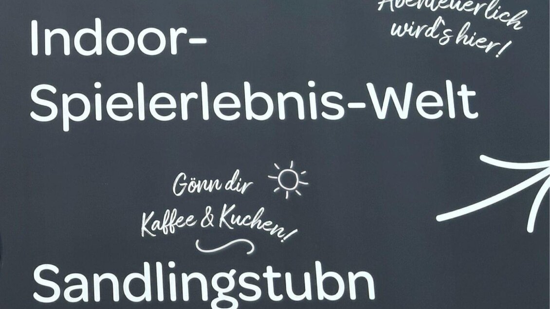 Ein Schild mit dem Text "Indoor-Spielerlebnis-Welt" und "Sandlingstuben". Es lädt dazu ein, Kaffee und Kuchen zu genießen. | © JUFA Hotels