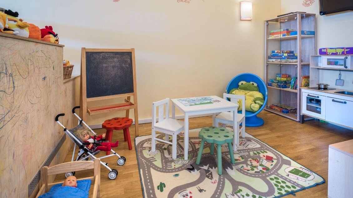 Ein helles Kinderzimmer mit einem Tisch, zwei Stühlen und einem Teppich mit Straßenmotiven. Es gibt Spielzeug, eine Tafel und eine gemütliche Sitzecke. | © JUFA Hotels