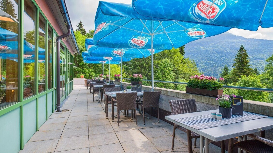 Eine einladende Terrasse mit Tischen und Stühlen, die von blauen Sonnenschirmen beschattet wird. Im Hintergrund sind Bäume und Berge sichtbar. | © JUFA Hotels
