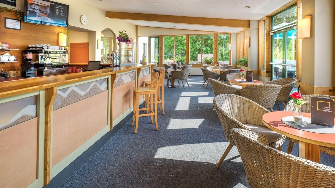 Ein gemütliches Café mit einem hellen, einladenden Ambiente und vielen Fenstern. Die Einrichtung besteht aus Rattanmöbeln und einer langen Theke. | © JUFA Hotels