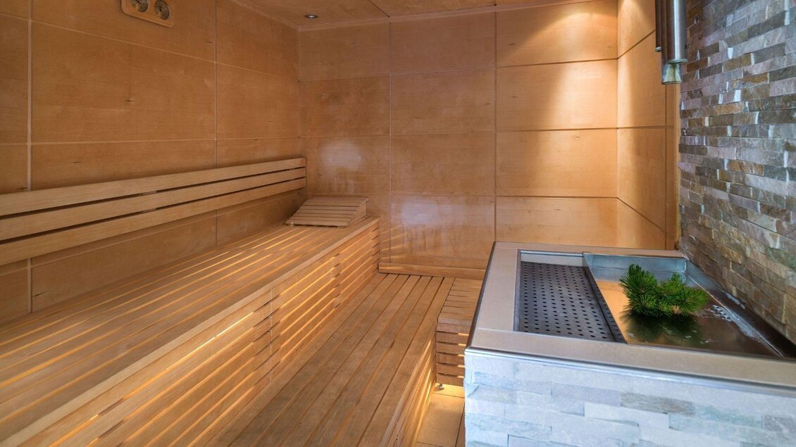 Eine moderne Sauna mit Holzlattenbänken und sanfter Beleuchtung. An der Wand befindet sich ein Steinbereich mit einem kleinen Pflanzenarrangement. | © JUFA Hotel