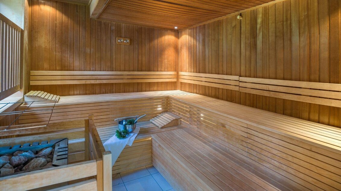 Eine gemütliche Sauna aus hellem Holz mit Bänken und einem kleinen Bereich für Saunazubehör. Im Hintergrund steht eine Schale mit Steinen und ein kleiner Tisch. | © JUFA Hotels