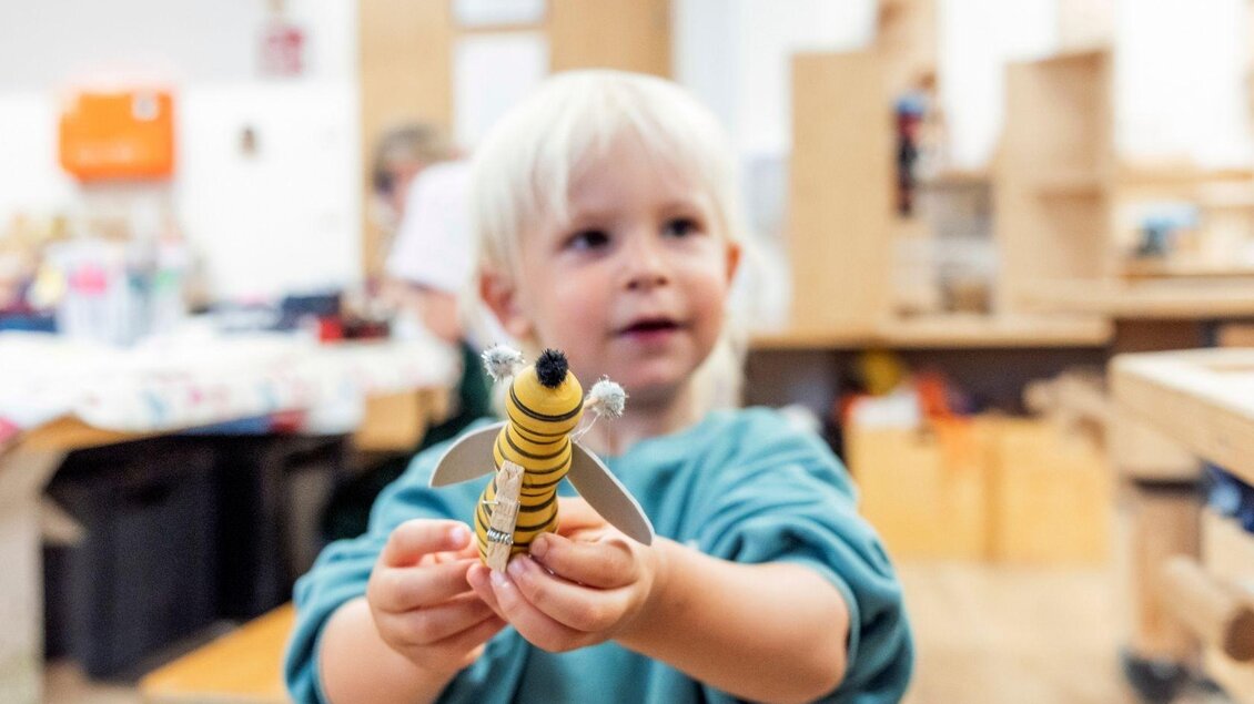 Ein kleines Kind hält stolz ein selbstgemachtes Spielzeug in den Händen. Im Hintergrund sind Werkbänke und weitere Kinder bei kreativen Aktivitäten zu sehen. | © www.zloam.at