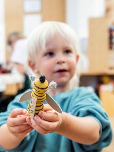Ein kleines Kind hält stolz ein selbstgemachtes Spielzeug in den Händen. Im Hintergrund sind Werkbänke und weitere Kinder bei kreativen Aktivitäten zu sehen. | © www.zloam.at