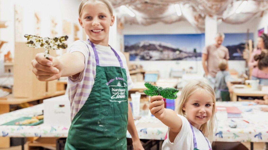 Zwei Mädchen halten stolz bunte Bastelarbeiten in den Händen. Im Hintergrund sieht man eine kreative Werkstatt mit mehreren Personen und Materialien. | © www.zloam.at