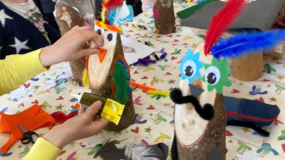Bunte Bastelprojekte aus Holz mit Federn und Augen. Kinder gestalten fröhliche Figuren auf einem Tisch mit buntem Papier und Bastelmaterialien. | © Narzissendorf Zloam