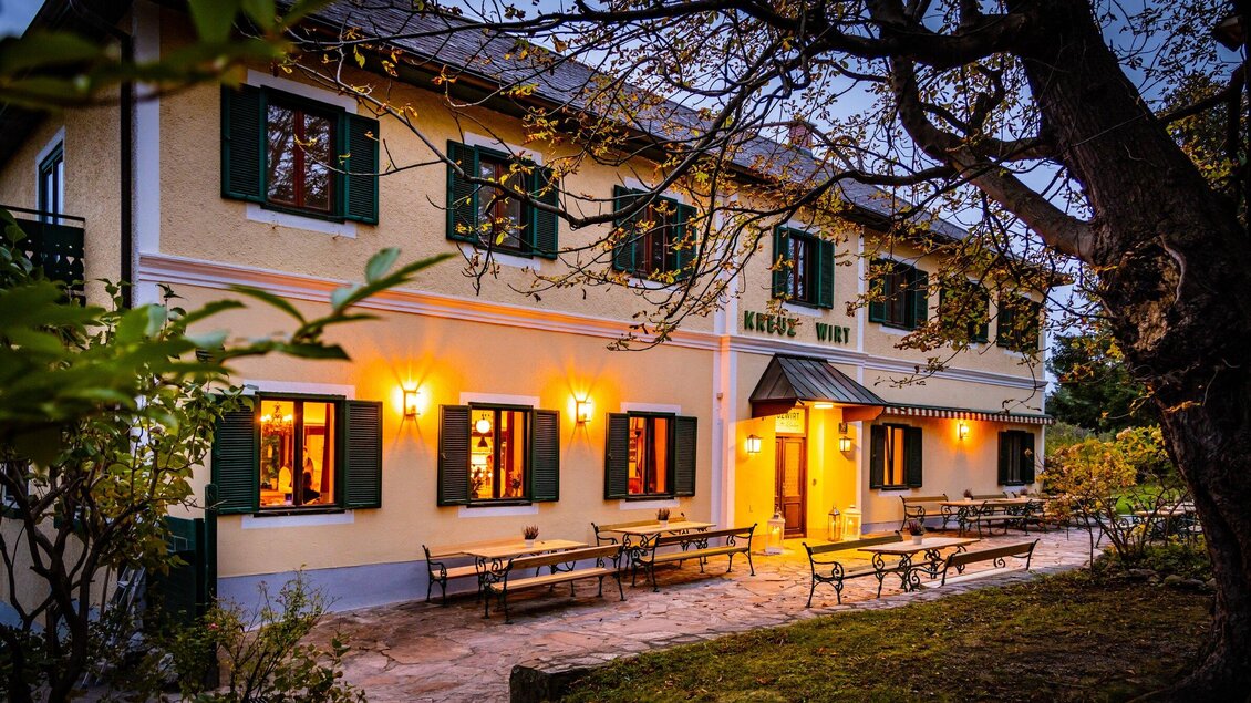 Ein charmantes Gasthaus mit warm beleuchteten Fenstern und einem einladenden Eingang. Der Garten bietet Sitzgelegenheiten im Freien unter Bäumen. | © Kreuzwirt am Rosenberg - Werner Krug