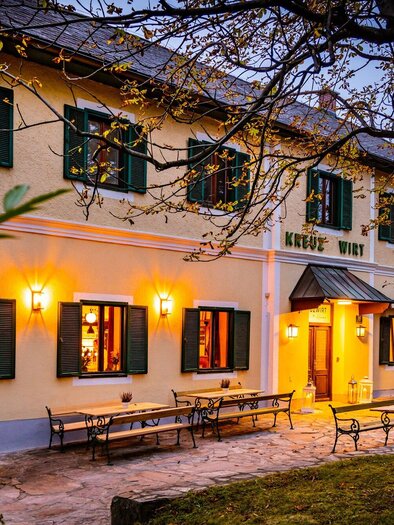A charming inn with warmly lit windows and an inviting entrance. The garden offers outdoor seating under trees. | © Kreuzwirt am Rosenberg - Werner Krug