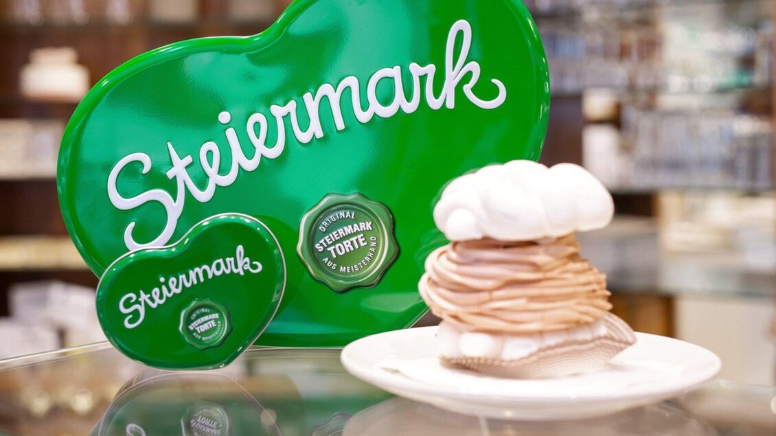 Ein Herz aus grünem Metall mit der Aufschrift „Steiermark“ steht neben einer weißen Torte. Die Torte hat eine dekorative Sahnehaube und ist auf einem Teller präsentiert. | © Harald Tauderer und Michaela Begsteiger