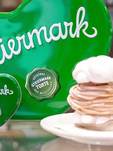 A heart made of green metal with the inscription "Styria" stands next to a white cake. The cake has a decorative cream topping and is presented on a plate. | © Harald Tauderer und Michaela Begsteiger