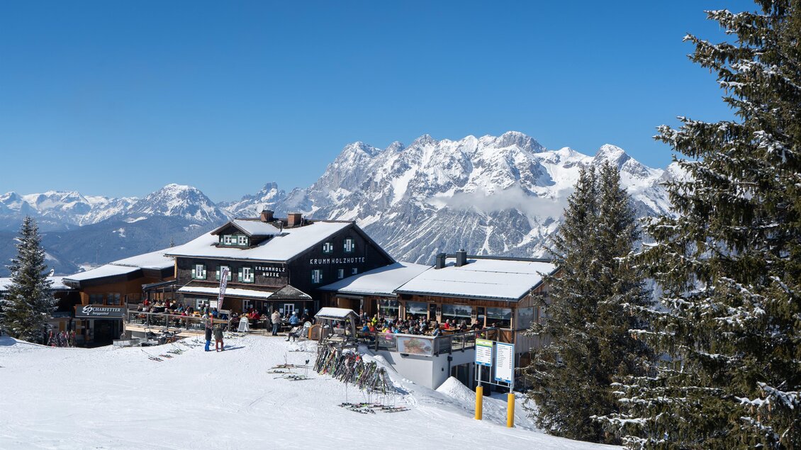 Außenansicht der Krummholzhütte am Hauser Kaibling mit Skifahrern und verschneiten Bergen im Hintergrund. | © Krummholzhütte