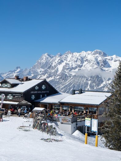 Exterior view of the Krummholz hut at Hauser Kaibling with skiers and snow-covered mountains in the background. | © Krummholzhütte