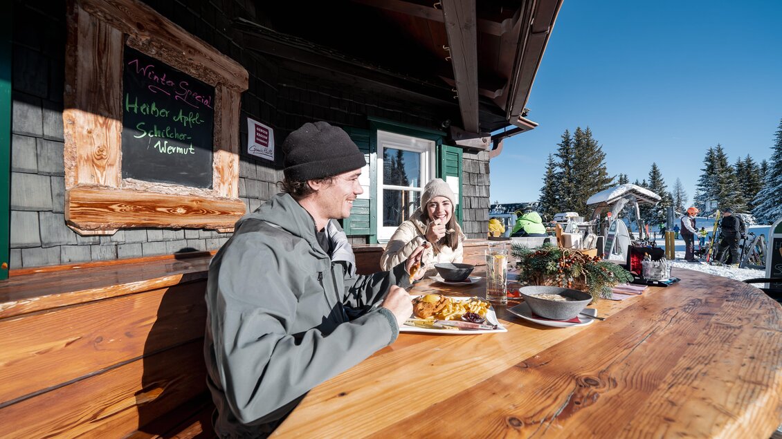 Zwei Gäste genießen ihre Mahlzeit an einem rustikalen Holztisch im Freien vor der Krummholzhütte. | © Krummholzhütte
