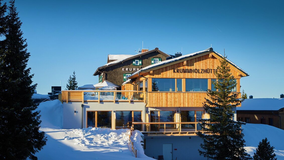 Ein gemütliches Berghaus aus Holz inmitten von Schnee. Der klare Himmel und die umliegenden Bäume verleihen der Szenerie eine ruhige Atmosphäre. | © Krummholzhütte