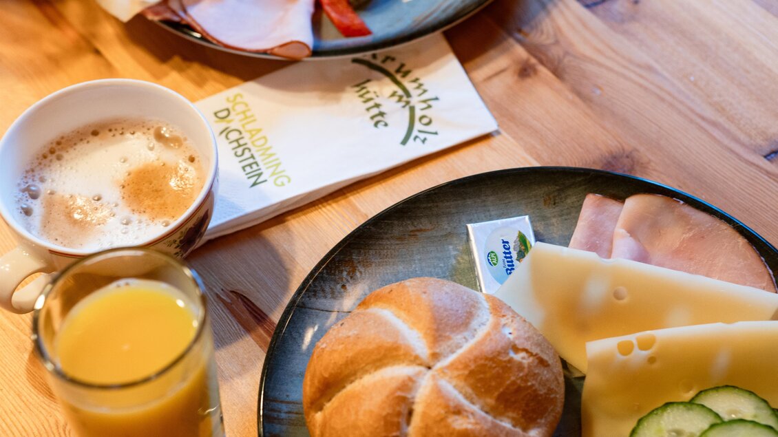 Frühstück mit Semmel, Käse, Schinken und Gurken sowie Getränk stehen am Tisch in der Krummholzhütte am Hauser Kaibling. Außerdem sieht man eine Tasse Kaffee und eine Serviette. | © Krummholzhütte