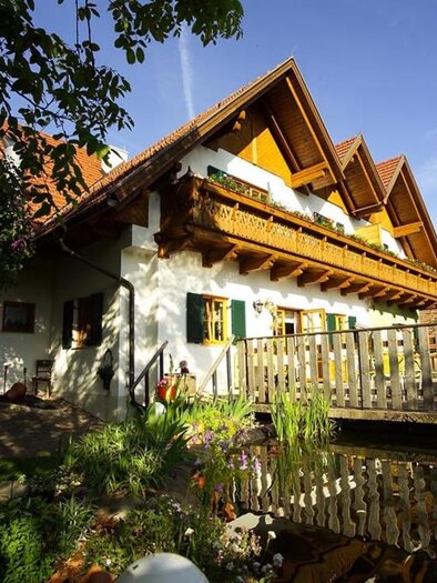Ein charmantes altes Bauernhaus mit einem Balkon und Holzverzierung. Umgeben von grünen Pflanzen und einem kleinen Teich. | © Kulinarischer Weinhof Florlwirt